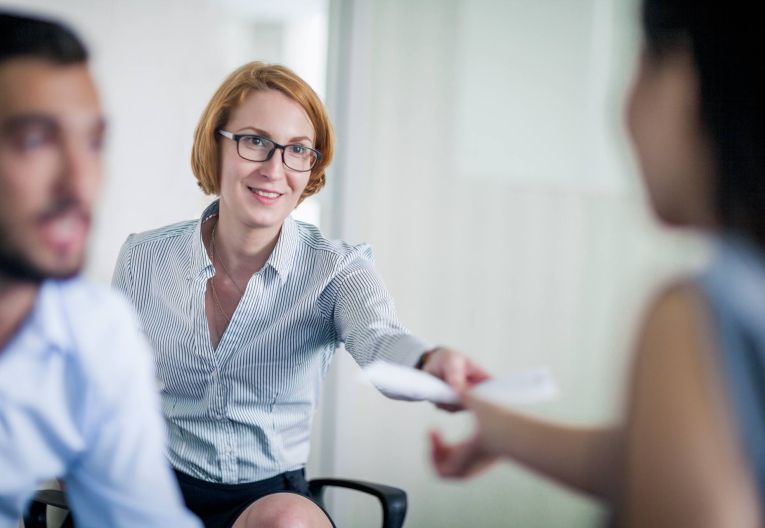 Frau mit kurzen Haaren und Brille übergibt ein Dokument an eine andere Person in einem Büro.